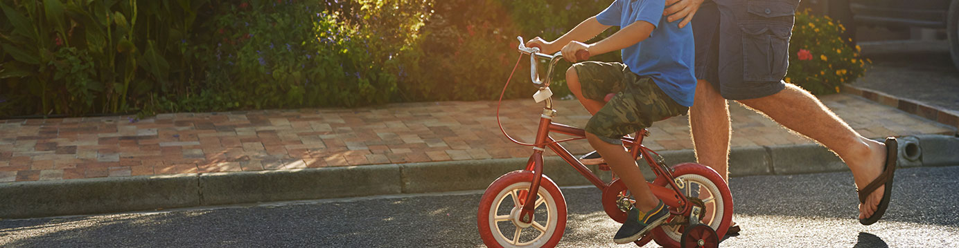 Child riding a bike with adult walking alongside on neighborhood street.